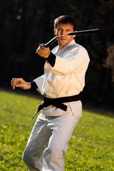 Kung fu expert in white kimono with black belt is practicing with nunchaku outdoors.