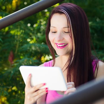 Smiling woman is using her white digital tablet outdoors in nature on green background.