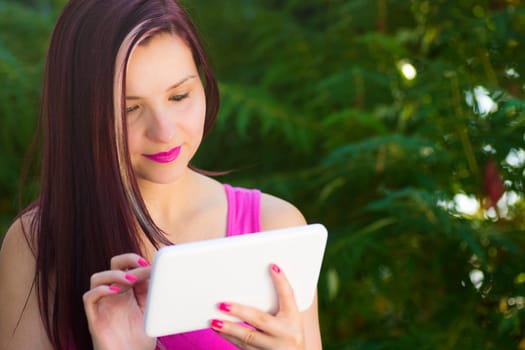 Smiling woman is using her white digital tablet outdoors in nature on green background.