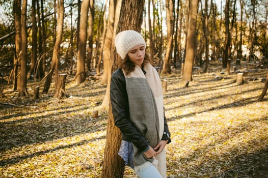 young woman walks among the trees on the bank of a sunny day. dressed in a black leather jacket, blue jeans, brown boots, a hat and scarf, autumn and spring outfit