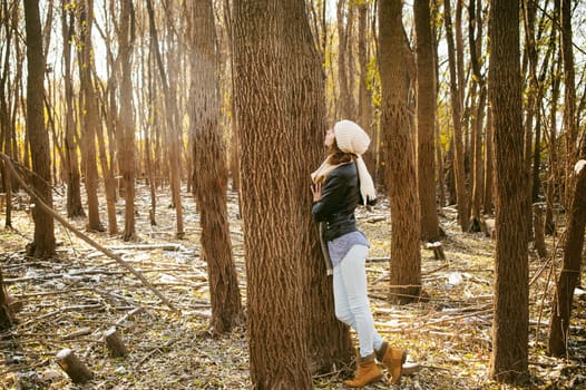 young woman walks among the trees on the bank of a sunny day. dressed in a black leather jacket, blue jeans, brown boots, a hat and scarf, autumn and spring outfit