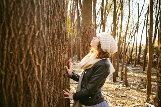 young woman walks among the trees on the bank of a sunny day. dressed in a black leather jacket, blue jeans, brown boots, a hat and scarf, autumn and spring outfit