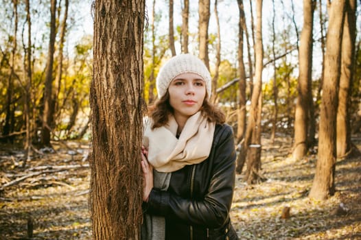 young woman walks among the trees on the bank of a sunny day. dressed in a black leather jacket, blue jeans, brown boots, a hat and scarf, autumn and spring outfit