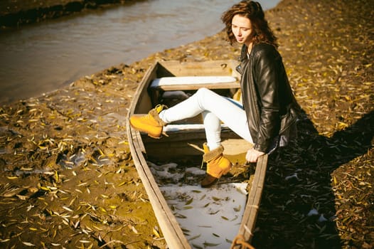 Slim girl in blue jeans, brown jacket and boots, sat on the upturned boat, on the shore, yellow autumn leaves bright sunny day