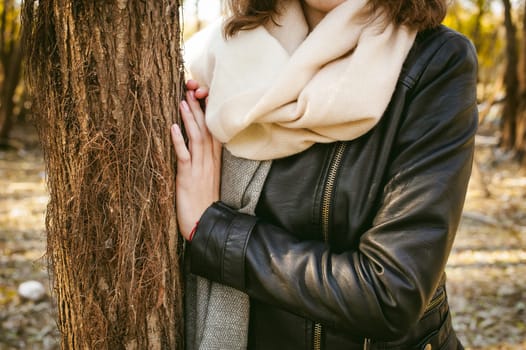 young woman walks among the trees on the bank of a sunny day. dressed in a black leather jacket, blue jeans, brown boots, a hat and scarf, autumn and spring outfit