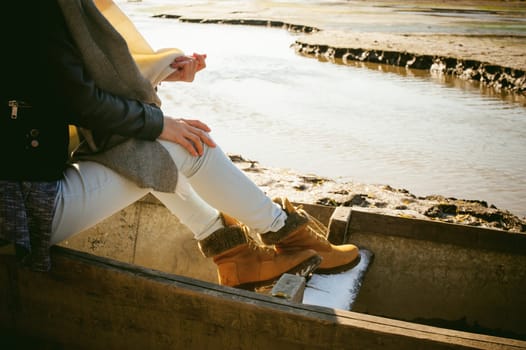 slim girl's legs in blue jeans and brown boots. girl sat on the upturned boat, on the shore, yellow autumn leaves