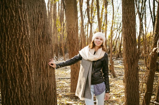 young woman walks among the trees on the bank of a sunny day. dressed in a black leather jacket, blue jeans, brown boots, a hat and scarf, autumn and spring outfit