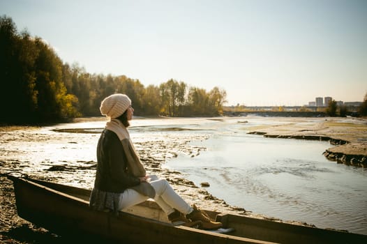 Slim girl in blue jeans, brown jacket and boots, sat on the upturned boat, on the shore, yellow autumn leaves bright sunny day