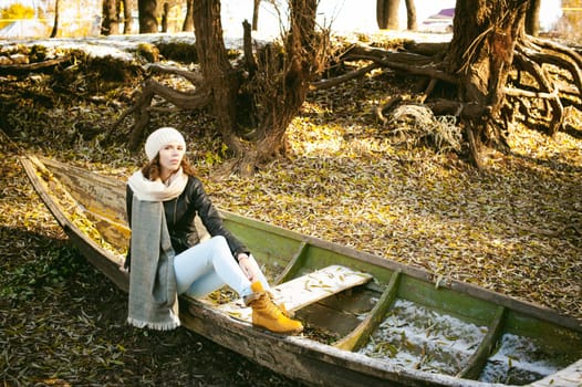 Slim girl in blue jeans, brown jacket and boots, sat on the upturned boat, on the shore, yellow autumn leaves bright sunny day