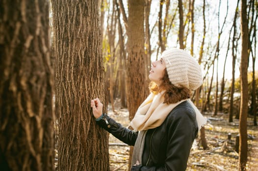 young woman walks among the trees on the bank of a sunny day. dressed in a black leather jacket, blue jeans, brown boots, a hat and scarf, autumn and spring outfit