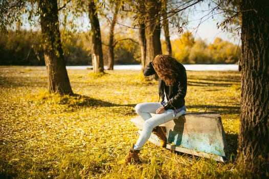 Slim girl in blue jeans, brown jacket and boots, sat on the upturned boat, on the shore, yellow autumn leaves bright sunny day