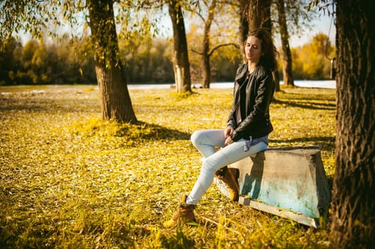 Slim girl in blue jeans, brown jacket and boots, sat on the upturned boat, on the shore, yellow autumn leaves bright sunny day