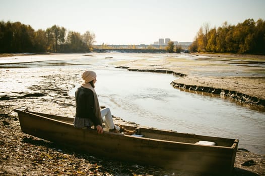 Slim girl in blue jeans, brown jacket and boots, sat on the upturned boat, on the shore, yellow autumn leaves bright sunny day