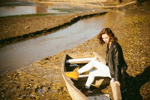 Slim girl in blue jeans, brown jacket and boots, sat on the upturned boat, on the shore, yellow autumn leaves bright sunny day
