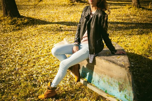 Slim girl in blue jeans, brown jacket and boots, sat on the upturned boat, on the shore, yellow autumn leaves bright sunny day