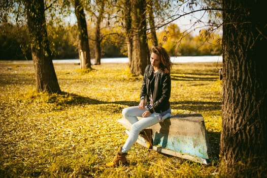 Slim girl in blue jeans, brown jacket and boots, sat on the upturned boat, on the shore, yellow autumn leaves bright sunny day