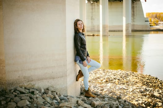 young woman in blue jeans and jacket, standing on the river bank with his back to the support of the bridge bent knee