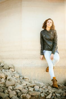 beautiful legs of the young girl in blue jeans, a leather jacket and brown boots. woman standing his back against the concrete wall, the stones beneath her feet. lit by beautiful warm autumn sunshine