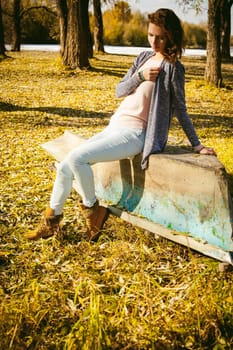 Slim girl in blue jeans, brown jacket and boots, sat on the upturned boat, on the shore, yellow autumn leaves bright sunny day