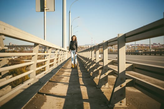 girl goes on road bridge. lonely young woman in black Leather jacket autumn, walking over the bridge which goes transport.