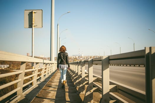 girl goes on road bridge. lonely young woman in black Leather jacket autumn, walking over the bridge which goes transport.