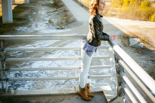 portrait of a beautiful young woman with long thin hair, a leather jacket, blue jeans. warm autumn sunny day on the stairs of the bridge.