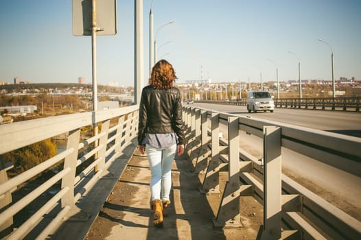 girl goes on road bridge. lonely young woman in black Leather jacket autumn, walking over the bridge which goes transport.