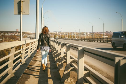 girl goes on road bridge. lonely young woman in black Leather jacket autumn, walking over the bridge which goes transport.