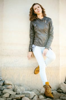 beautiful legs of the young girl in blue jeans, a leather jacket and brown boots. woman standing his back against the concrete wall, the stones beneath her feet. lit by beautiful warm autumn sunshine
