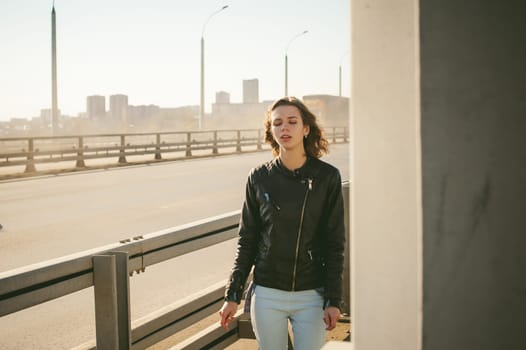 girl goes on road bridge. lonely young woman in black Leather jacket autumn, walking over the bridge which goes transport.