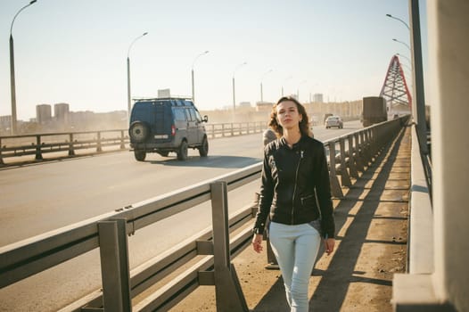 girl goes on road bridge. lonely young woman in black Leather jacket autumn, walking over the bridge which goes transport.