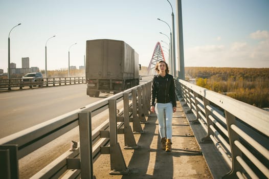 girl goes on road bridge. lonely young woman in black Leather jacket autumn, walking over the bridge which goes transport.