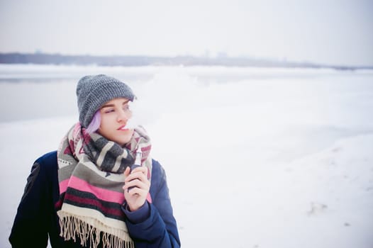 vaping girl. winter street portrait of a woman hipster, purple-dyed hair, a gray knitted hat and scarf. woman smokes an electronic cigarette in the street near the snow-covered river