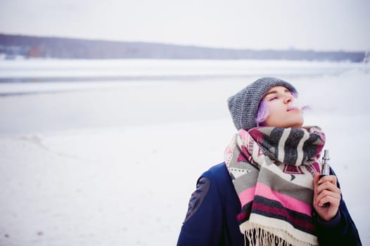 vaping girl. winter street portrait of a woman hipster, purple-dyed hair, a gray knitted hat and scarf. woman smokes an electronic cigarette in the street near the snow-covered river