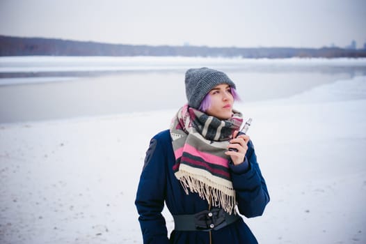 vaping girl. winter street portrait of a woman hipster, purple-dyed hair, a gray knitted hat and scarf. woman smokes an electronic cigarette in the street near the snow-covered river