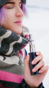 vaping girl. winter street portrait of a woman hipster, purple-dyed hair, a gray knitted hat and scarf. woman smokes an electronic cigarette in the street near the snow-covered river