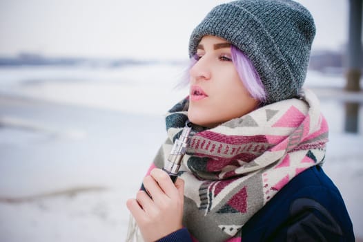 vaping girl. winter street portrait of a woman hipster, purple-dyed hair, a gray knitted hat and scarf. woman smokes an electronic cigarette in the street near the snow-covered river
