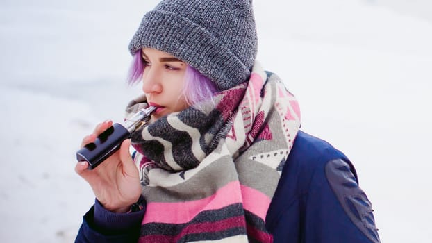 vaping girl. winter street portrait of a woman hipster, purple-dyed hair, a gray knitted hat and scarf. woman smokes an electronic cigarette in the street near the snow-covered river