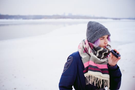vaping girl. winter street portrait of a woman hipster, purple-dyed hair, a gray knitted hat and scarf. woman smokes an electronic cigarette in the street near the snow-covered river