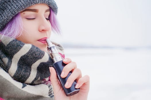 vaping girl. winter street portrait of a woman hipster, purple-dyed hair, a gray knitted hat and scarf. woman smokes an electronic cigarette in the street near the snow-covered river