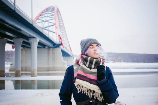 vaping girl. winter street portrait of a woman hipster, purple-dyed hair, a gray knitted hat and scarf. woman smokes an electronic cigarette in the street near the snow-covered river
