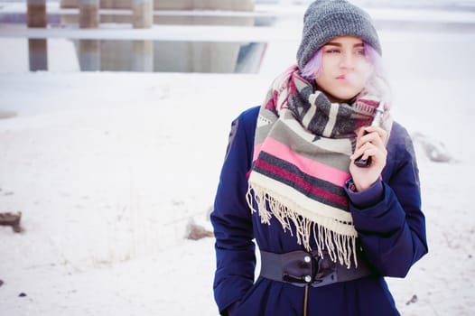 vaping girl. winter street portrait of a woman hipster, purple-dyed hair, a gray knitted hat and scarf. woman smokes an electronic cigarette in the street near the snow-covered river