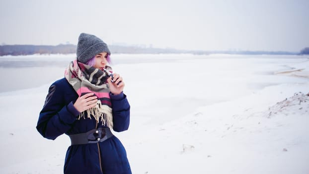 vaping girl. winter street portrait of a woman hipster, purple-dyed hair, a gray knitted hat and scarf. woman smokes an electronic cigarette in the street near the snow-covered river