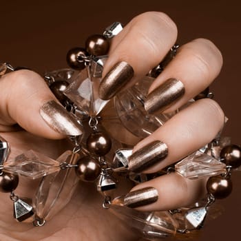 Female hand with shiny brown nails holds brown jewel on brown background.