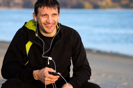 Smiling man with earphones is sitting outdoors along the riverside. Lifestyle concept.