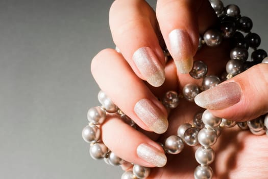 Female hand with shiny textured pearly nails is holding pearls on gray background.
