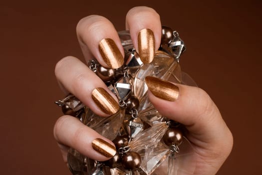 Female hand with shiny golden nails is holding brown jewel on brown background.