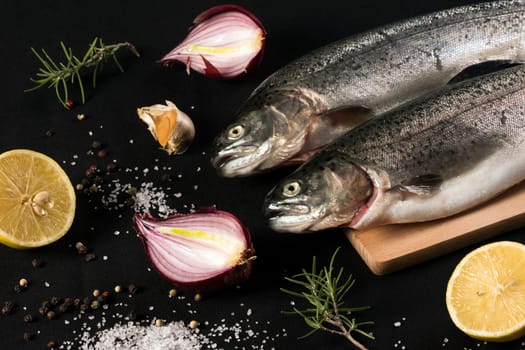 Raw trout fish with salt, pepper, lemon, garlic, onion and rosemary on black background.