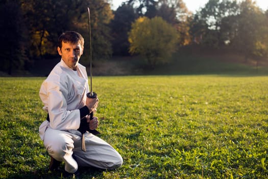 Ninja in white kimono with sword is outdoors in the park.
