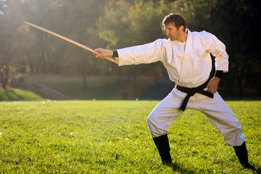 Man in white kimono is practicing with wooden sword outdoors in the park.